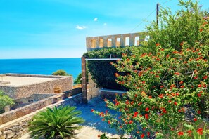 A stone pathway leading to a terrace with a view of the sea, surrounded by lush greenery and red flowers.