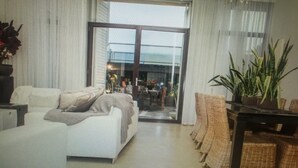 Una sala de estar moderna con un amplio ventanal corredera que da a un área exterior. Hay un sofá con cojines, una mesita con una planta y una silla de mimbre.