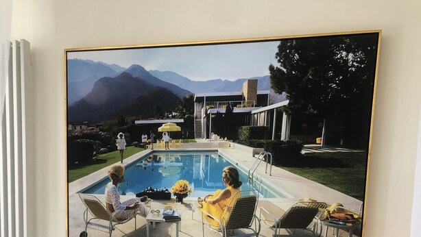 Un dormitorio con un cuadro grande que muestra una piscina y montañas, una cama con almohadas blancas y una lámpara de pie.