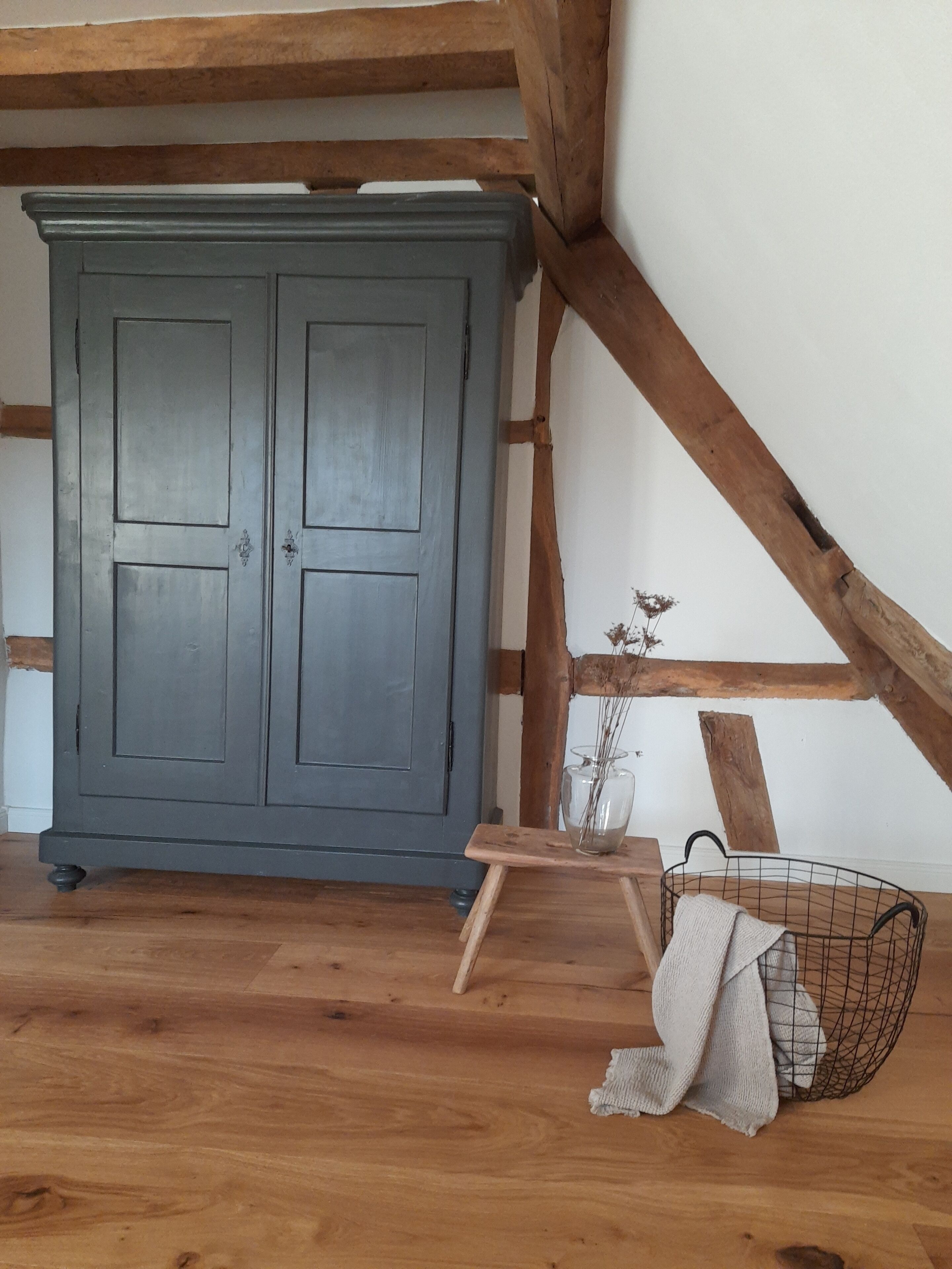 Ein Zimmer mit einem großen grauen Kleiderschrank, Holzbalken an der Decke, einem kleinen Holztisch mit einer Vase und einem Drahtkorb mit Stoff.