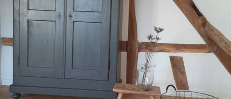 Ein Zimmer mit einem großen grauen Kleiderschrank, Holzbalken an der Decke, einem kleinen Holztisch mit einer Vase und einem Drahtkorb mit Stoff.