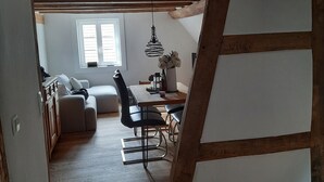 A loft-style room with exposed wooden beams, a dining area with a table and chairs, a sofa, and a window with blinds. - Modern vacation apartment "Tante Emma"  (Sinsheim)