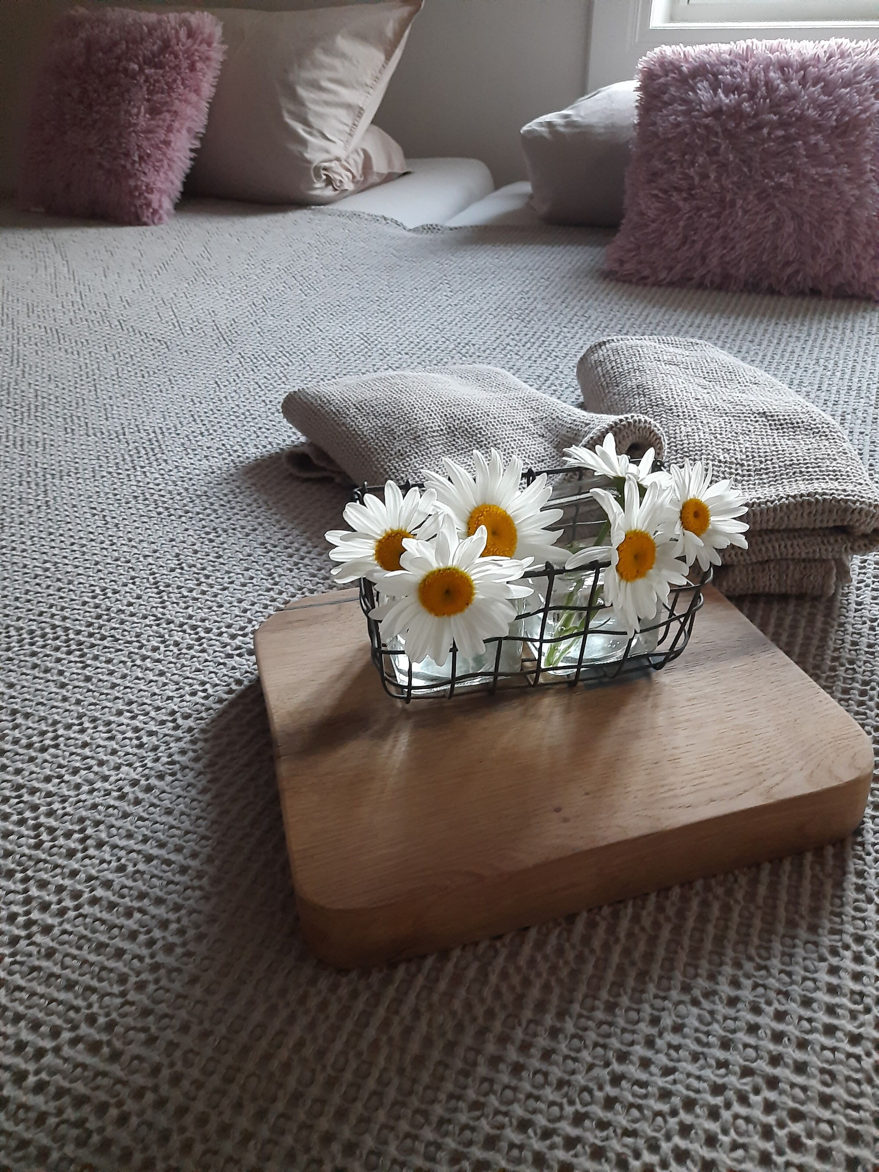 Un letto con una coperta grigia dalla trama decorativa, un comodino in legno con un cesto in filo che contiene fiori bianchi e una finestra dalla quale filtra la luce.