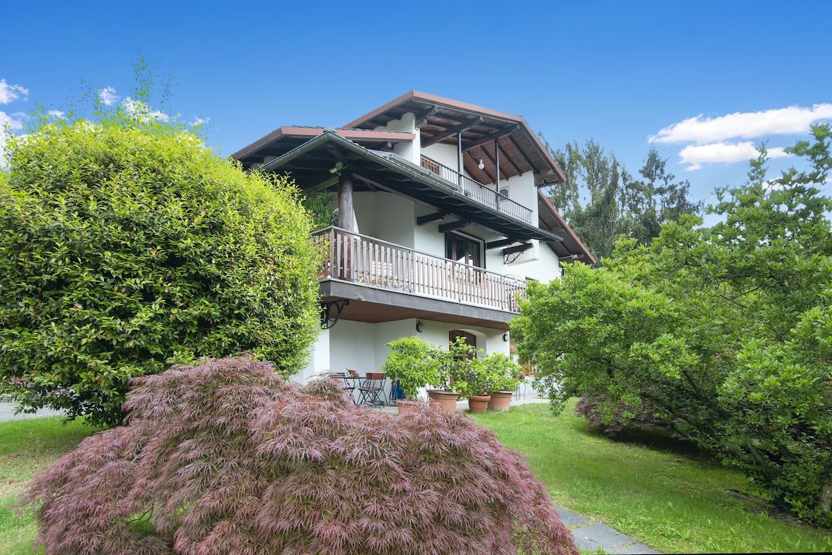 A two-story house with a balcony, surrounded by lush greenery and potted plants.