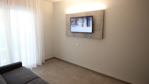 A modern hotel room with a flat-screen TV mounted on the wall, a grey sofa, and a large window with sheer curtains.