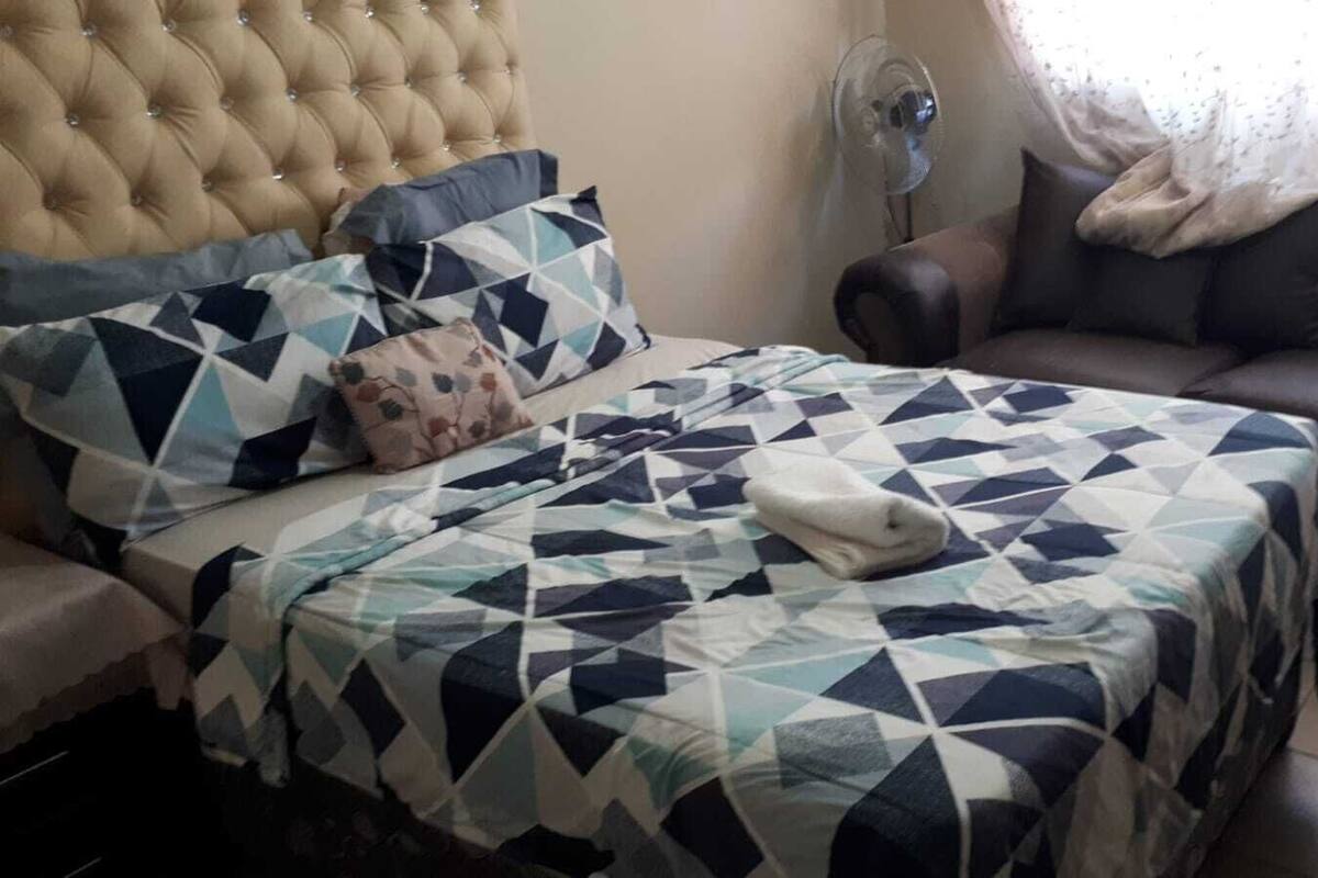 Ein Schlafzimmer mit einer gebügelten Kopfstütze, einer gesteppten Bettdecke, einem braunen Ledersofa und einem Standventilator.