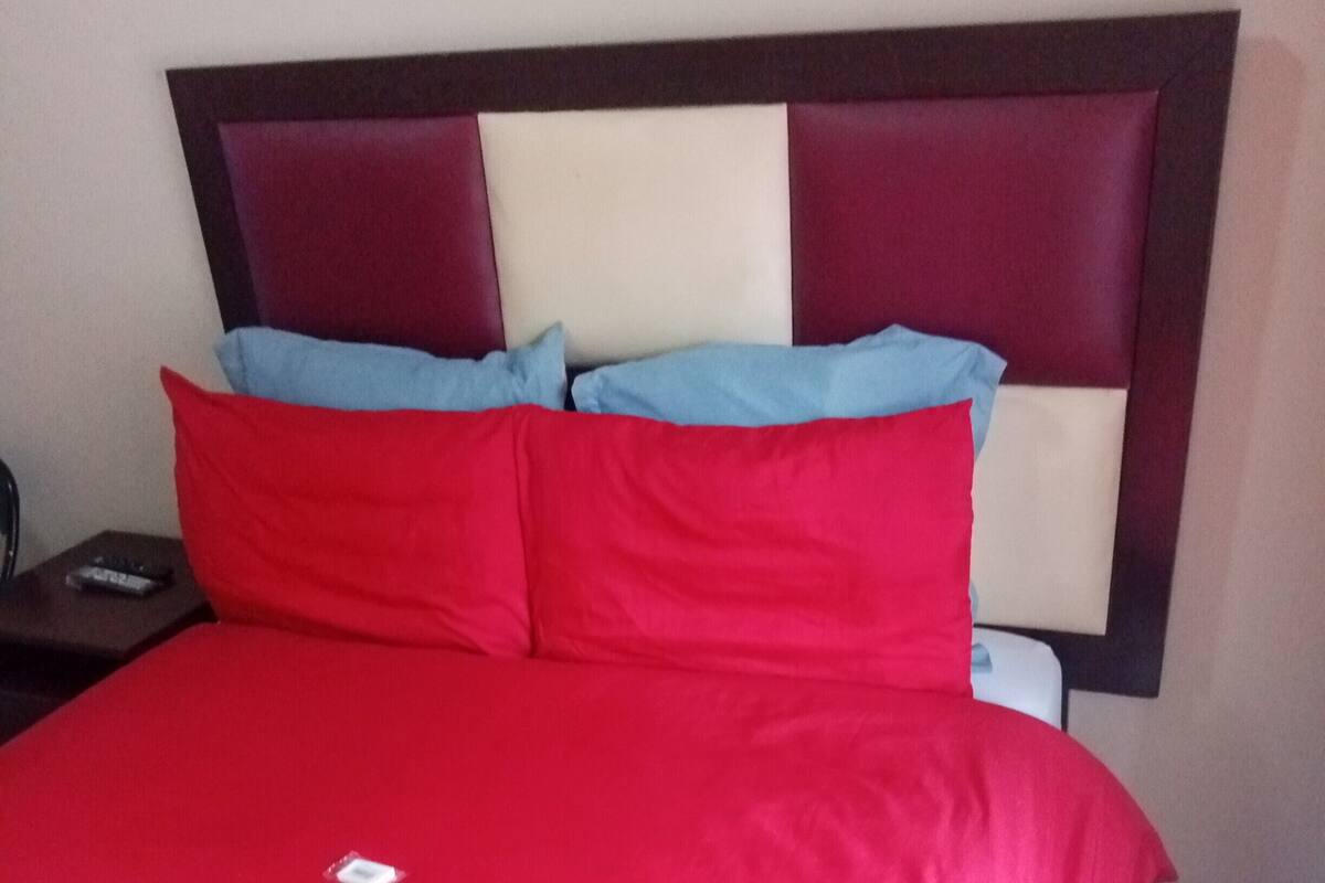 Ein Bett mit roter Bettdecke und zwei Kissen, ein Nachttisch mit Fernbedienung sowie eine Wand mit dunklem Kopfteil.