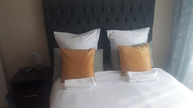 A neatly made bed with white linens and two decorative pillows, a dark headboard, and a small bedside table with a lamp. - C- Land Guest house Meyerton (Meyerton)