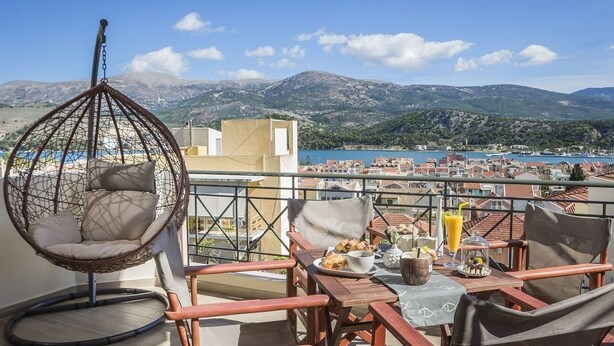 Un balcon avec un fauteuil suspendu, une table à manger avec des chaises, et une vue sur une ville côtière et des montagnes.