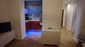 A modern living room with a flat-screen TV, a white sofa, and a small white table. A kitchen is visible in the background with red cabinets and a countertop. - Comfort and calm in the heart of Annecy (30m2 hyper center) (Annecy)