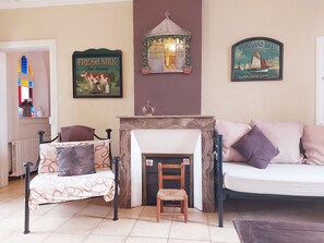 A cozy living room with a fireplace, a sofa, a chair, and a small table. There are two framed pictures on the wall, one of a cow and another of a ship. - Charming Farmhouse with Private Pool & Garden near Paris (La Chapelle-Fortin)