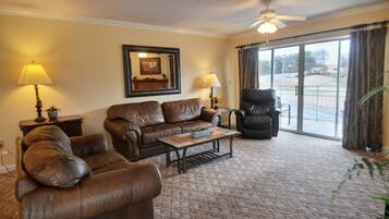 A living room with a brown leather sofa, a coffee table, and a chair. There is a large window with curtains, a ceiling fan, and a framed picture on the wall.