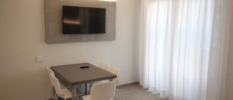 A modern room with a flat-screen TV mounted on the wall, a dining table with chairs, and sheer curtains on the window.