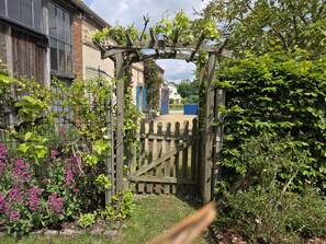 Une porte en bois mène à un jardin agrémenté d’une pergola, entouré de diverses plantes et d’une clôture.