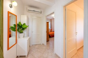 A hallway with a white cabinet, a potted plant, an air conditioning unit, and a door leading to a bedroom with a bed and a window.