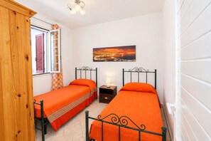 A small room with two single beds, a nightstand with a lamp, a window with shutters, and a wall-mounted picture.