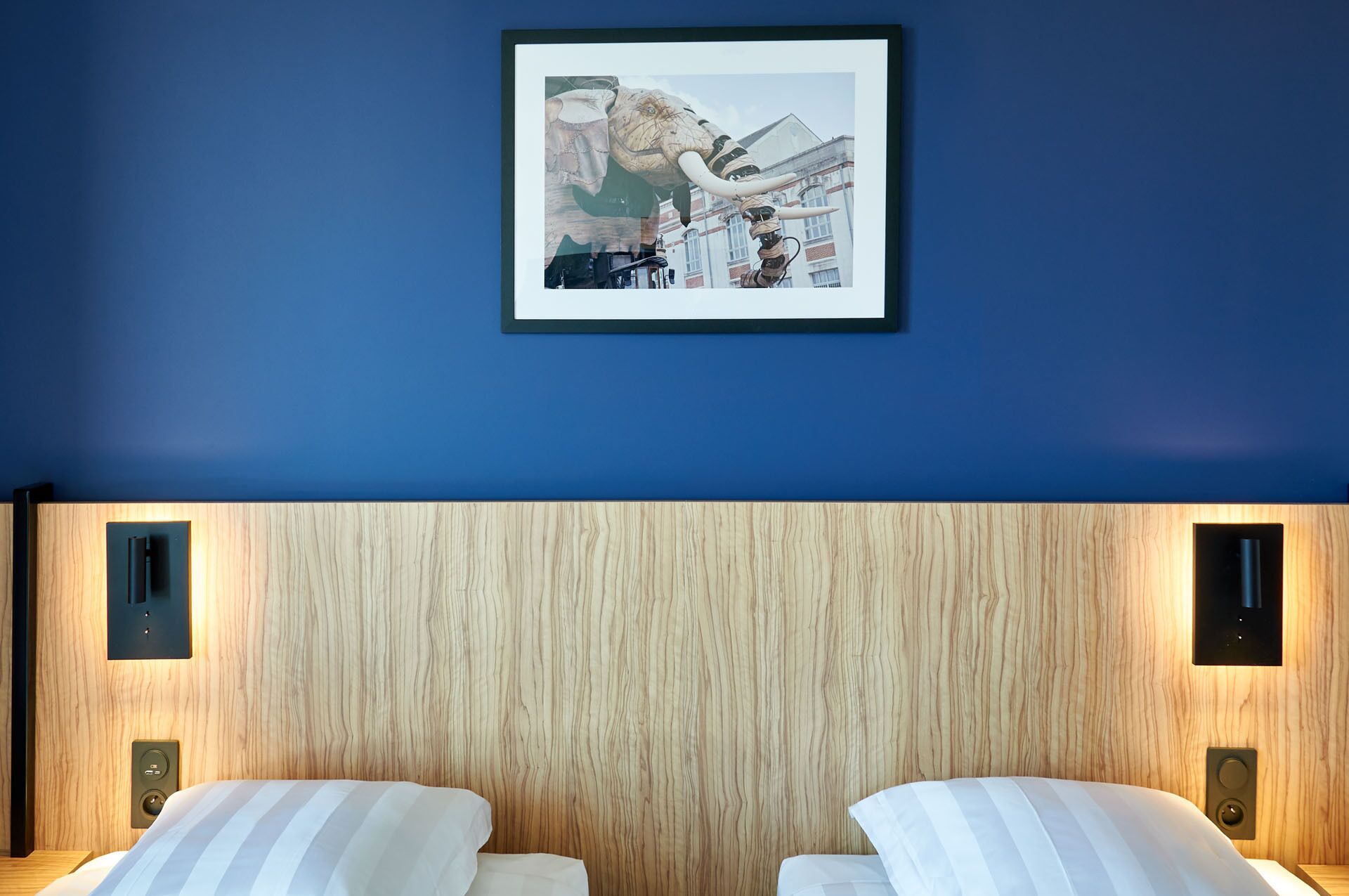 A hotel room with a blue wall, a framed picture of a mechanical elephant, and a wooden headboard.