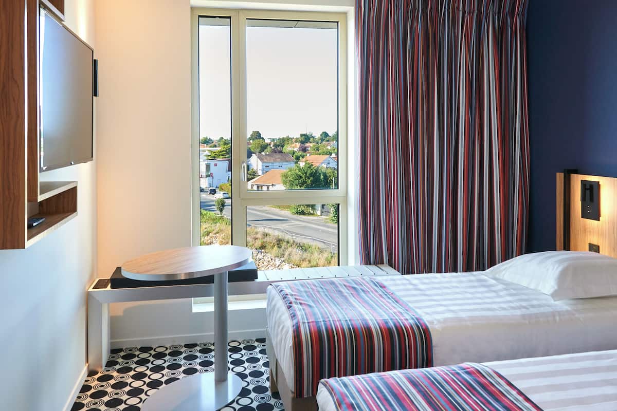 A hotel room with a large window, a bed, a small table, and a TV mounted on the wall.