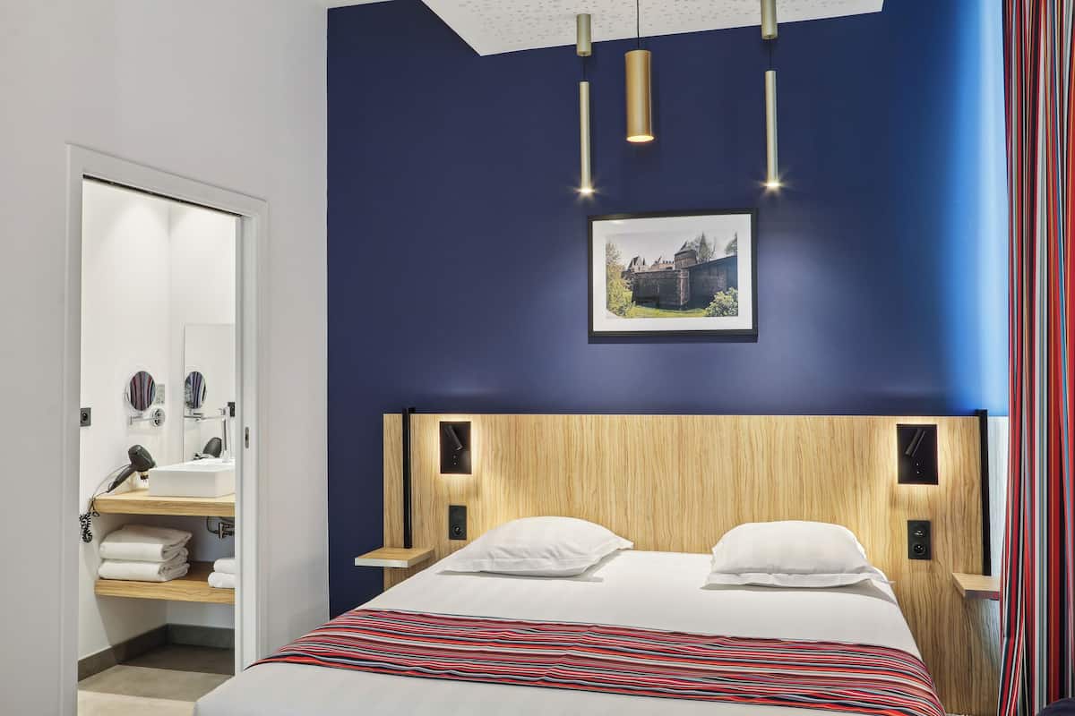 A modern bedroom with a wooden headboard, a bed with white linens, a bedside table with a lamp, and a framed picture on the wall.