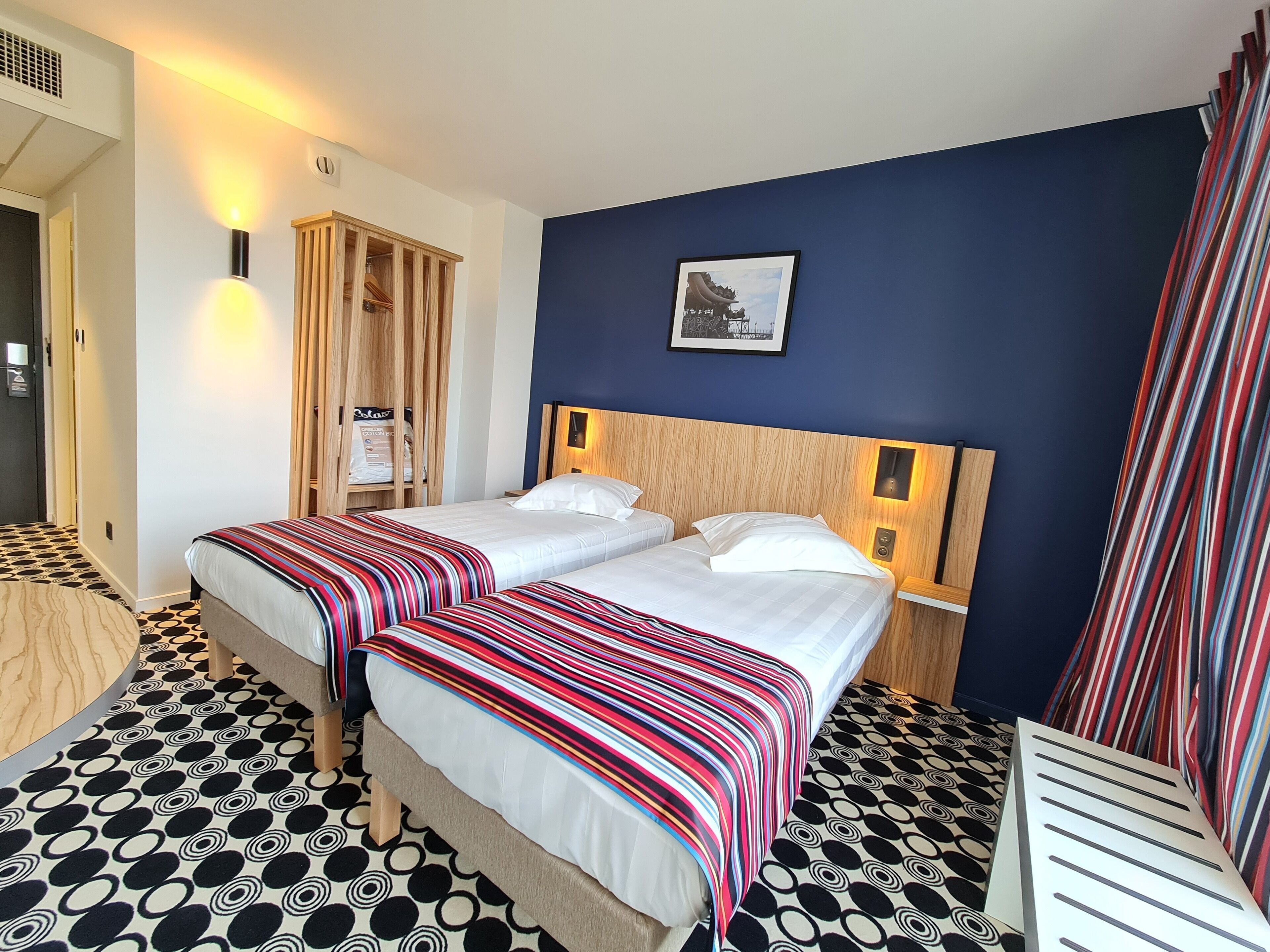 A hotel room with two beds, a wooden wardrobe, a framed picture on the wall, and a patterned floor.