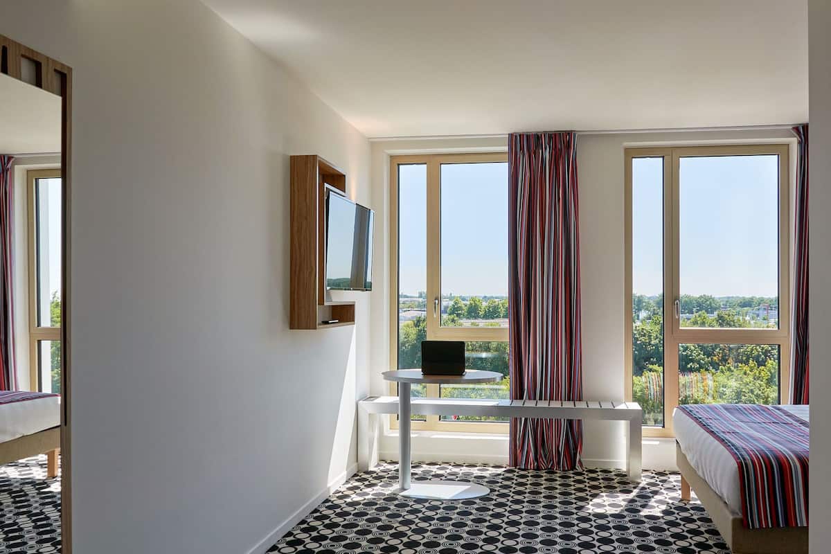 A hotel room with a bed, a desk, a TV mounted on the wall, and large windows with curtains.