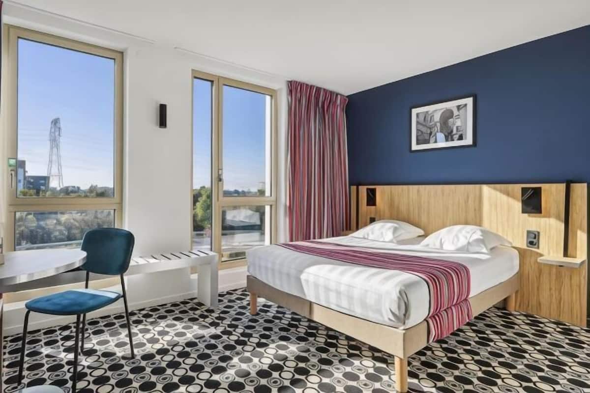 A hotel room with a bed, a desk, a chair, and large windows.