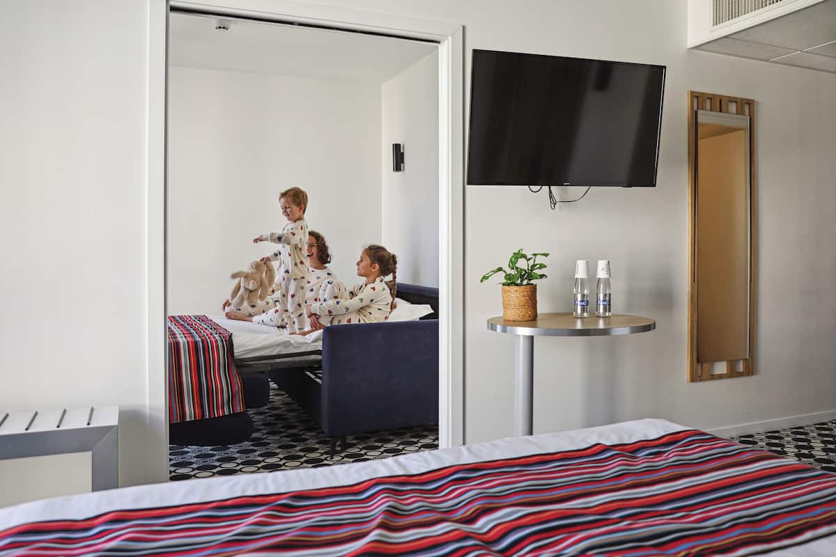 A hotel room with a bed, a TV mounted on the wall, a small table with a plant and bottles, and a mirror.