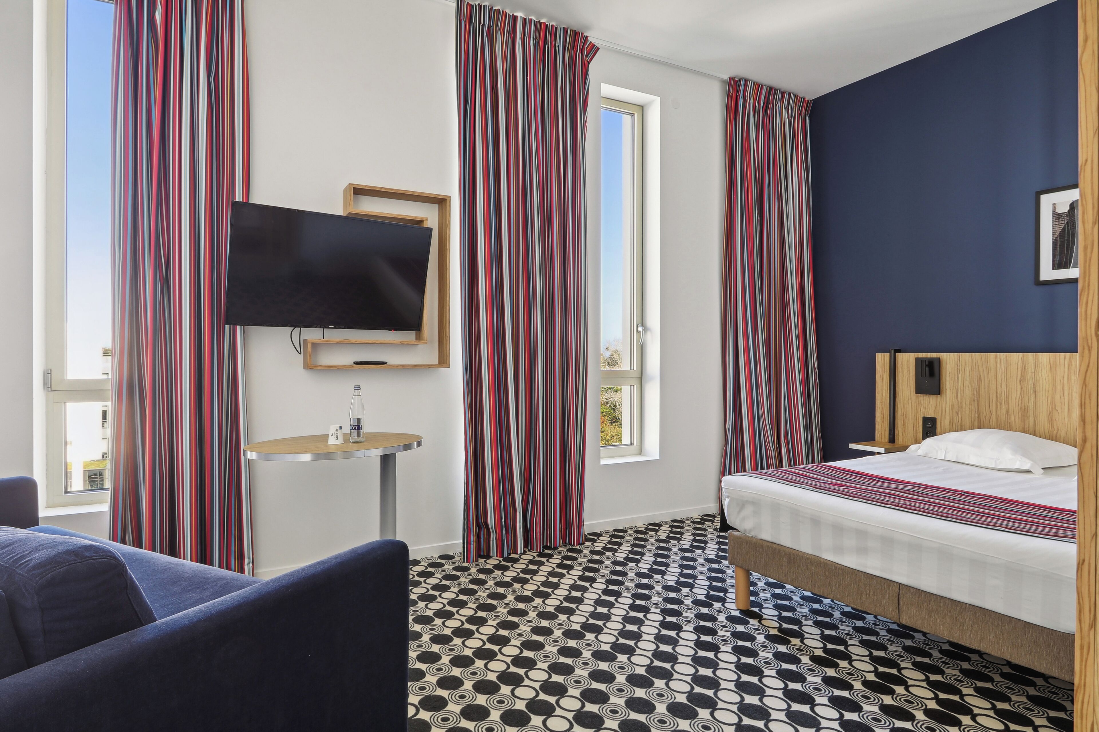 Una habitación de hotel con una cama, televisión, una mesita y una ventana con cortinas.