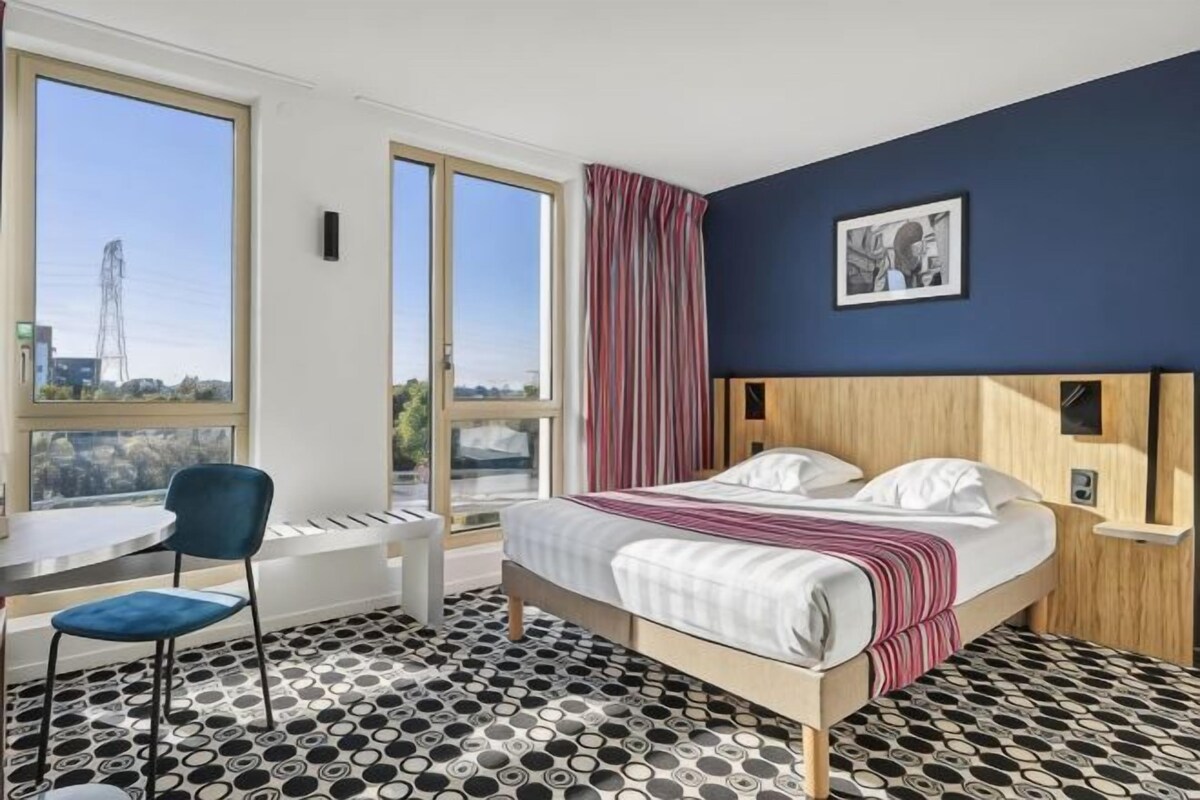 A hotel room with a bed, a desk, a chair, and large windows.