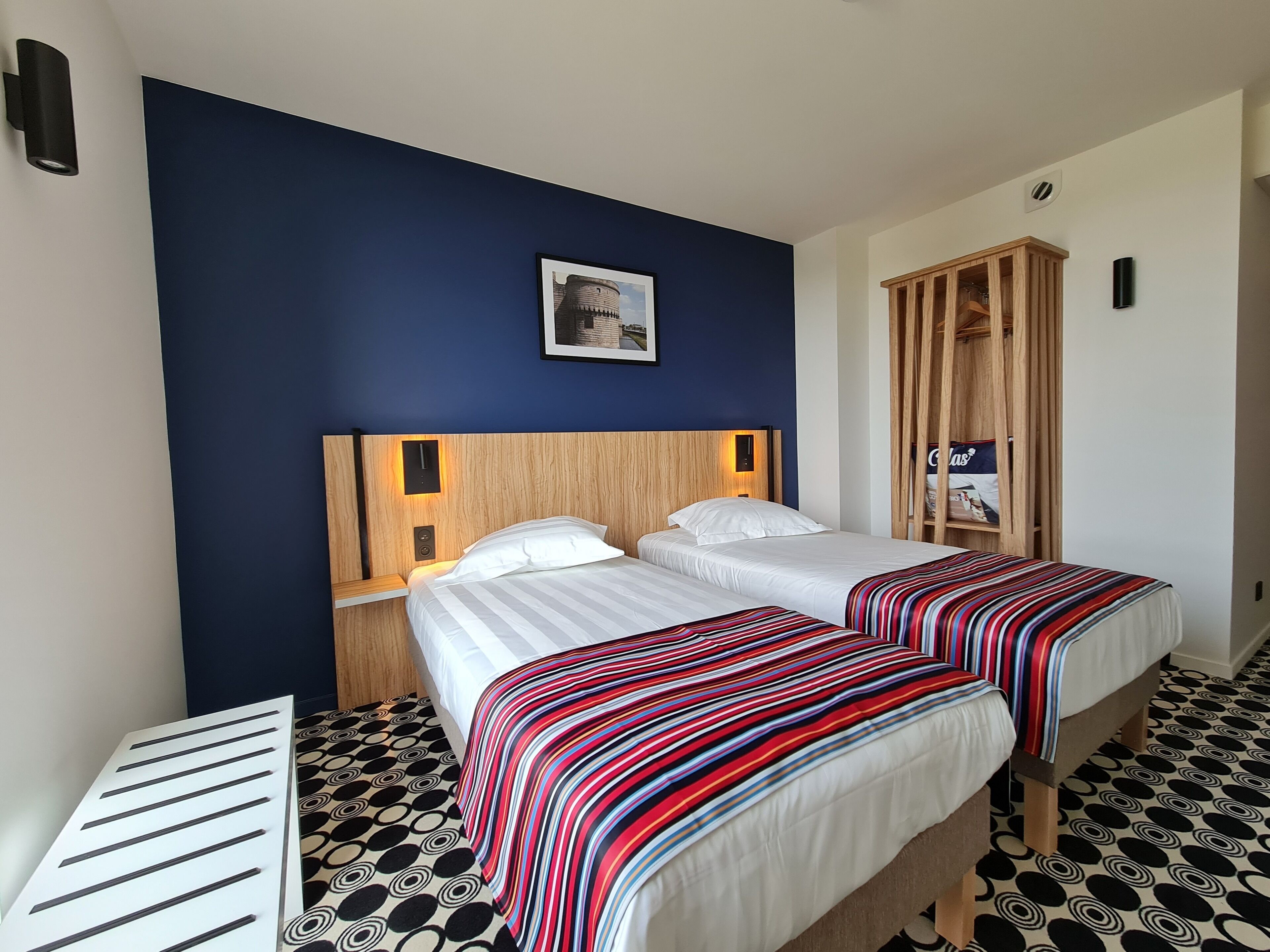 A hotel room with two beds, a wooden wardrobe, a framed picture on the wall, and a patterned floor.