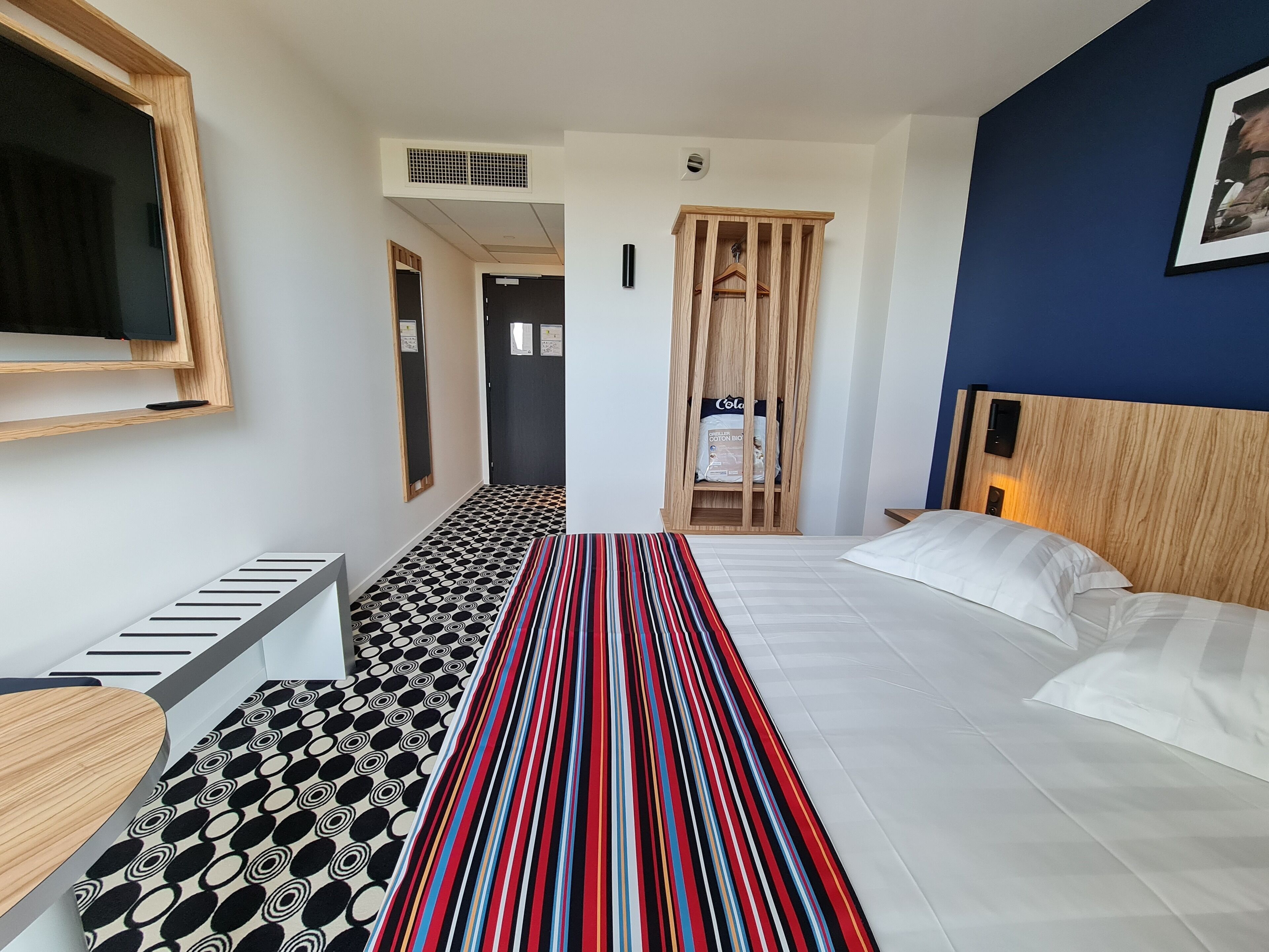 Une chambre d’hôtel avec un lit, un tapis à rayures, une armoire en bois, un téléviseur fixé au mur et un sol à motifs.