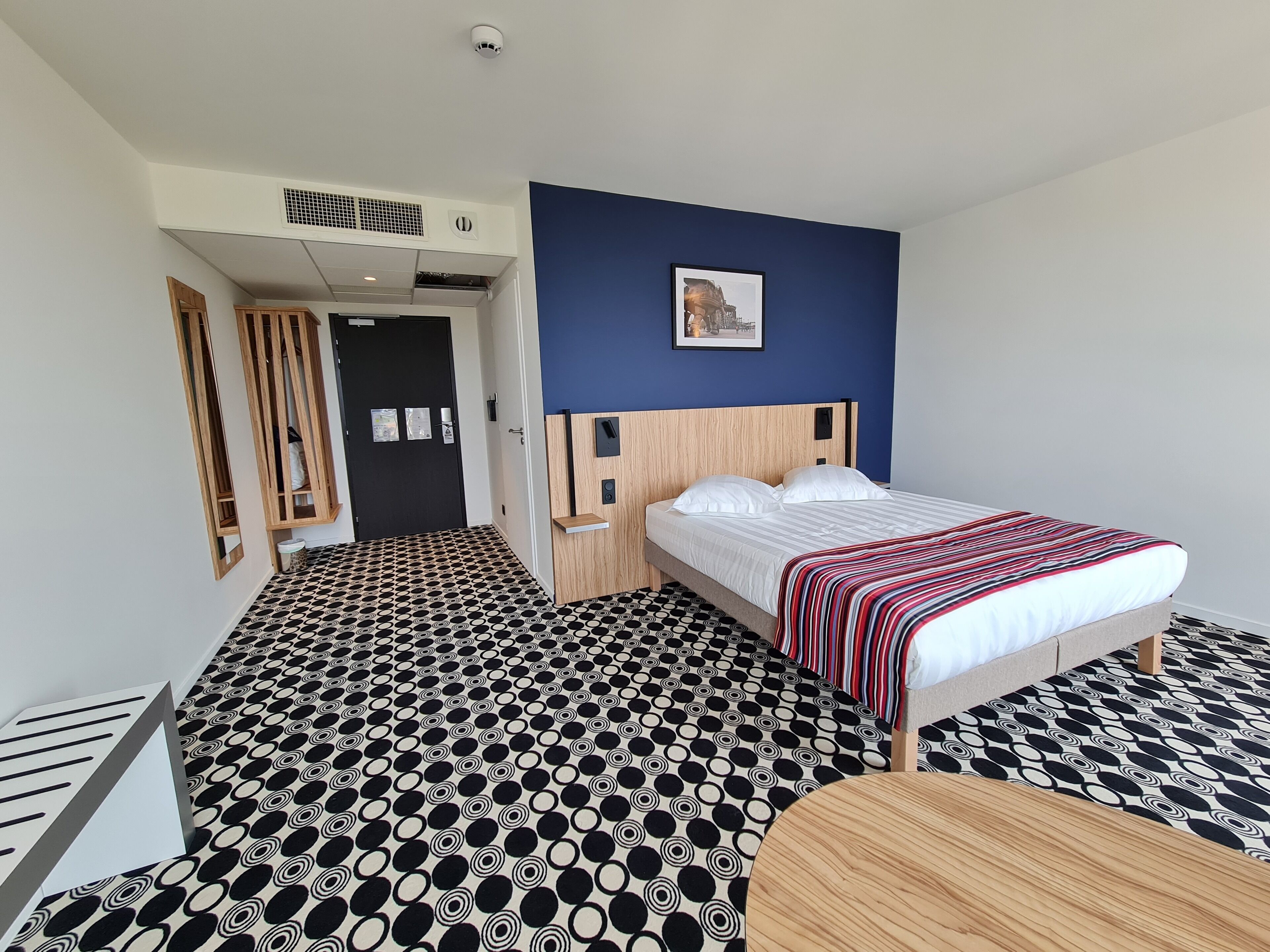 Une chambre d’hôtel avec un lit, une table de chevet, une petite table en bois, un sol à motifs, une porte et une bouche d’aération fixée au mur.