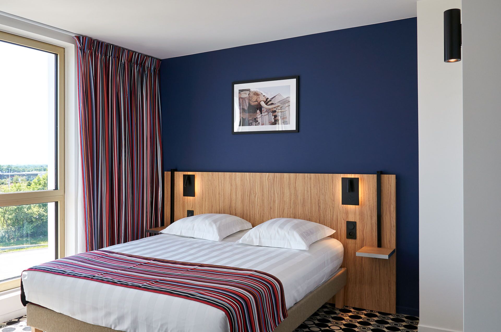 A bedroom with a large window, a bed with white bedding, a wooden headboard, and a framed picture on the wall.