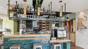 A bar area with a green-tiled bar counter, stainless steel stools, and a variety of bottles and glasses on shelves.