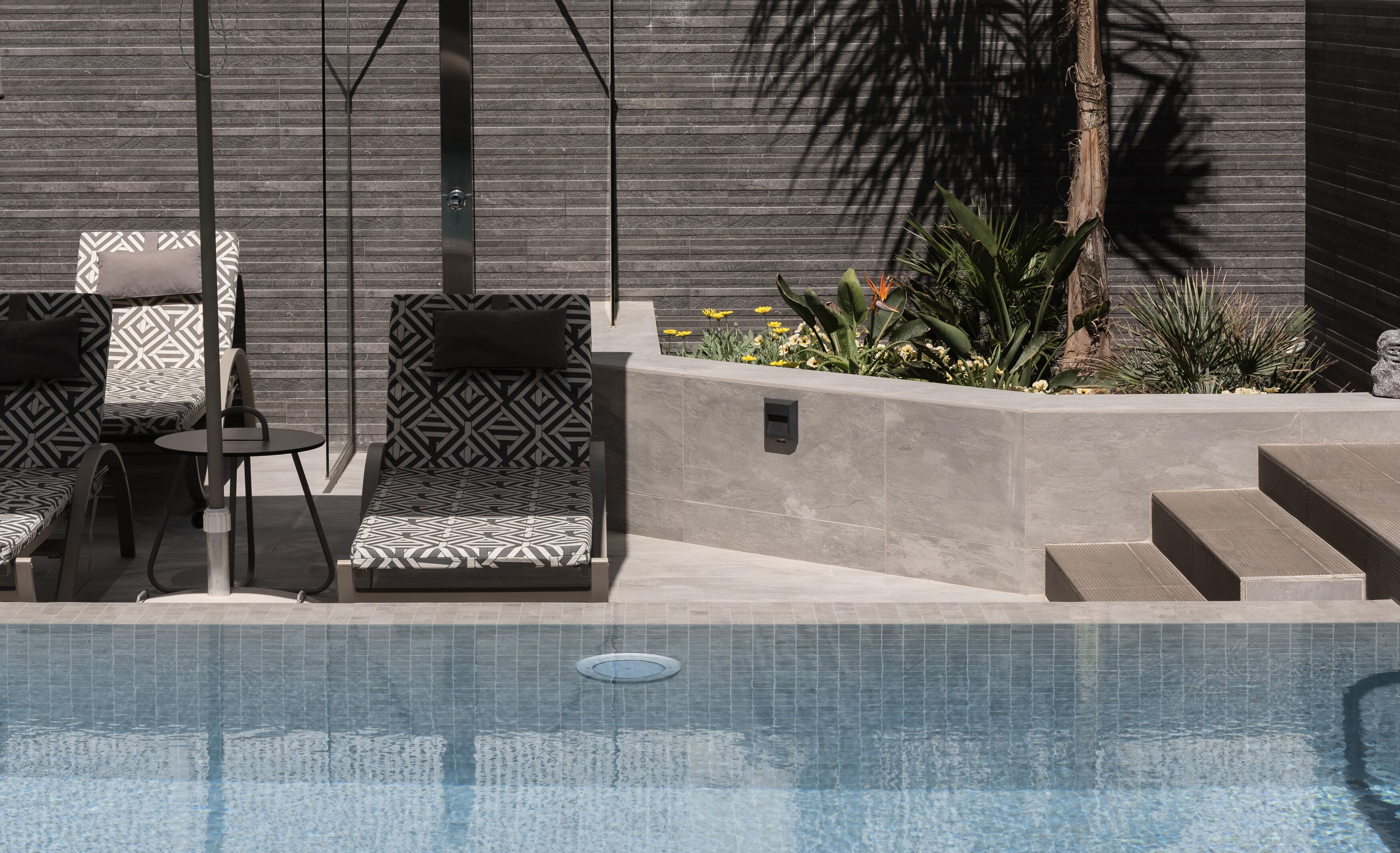 A modern outdoor pool area with geometric-patterned lounge chairs and a built-in planter with tropical plants.