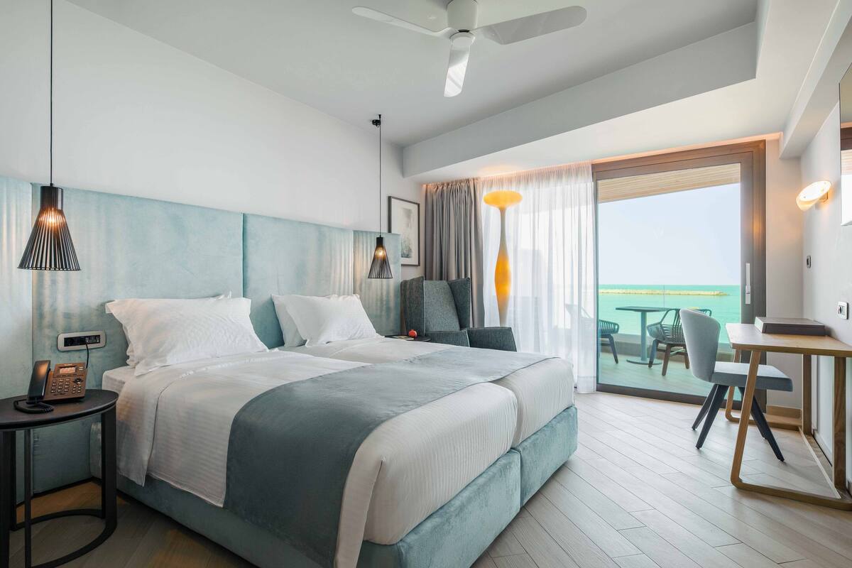 A modern hotel room with a large bed, a desk, and a balcony with a view of the sea.