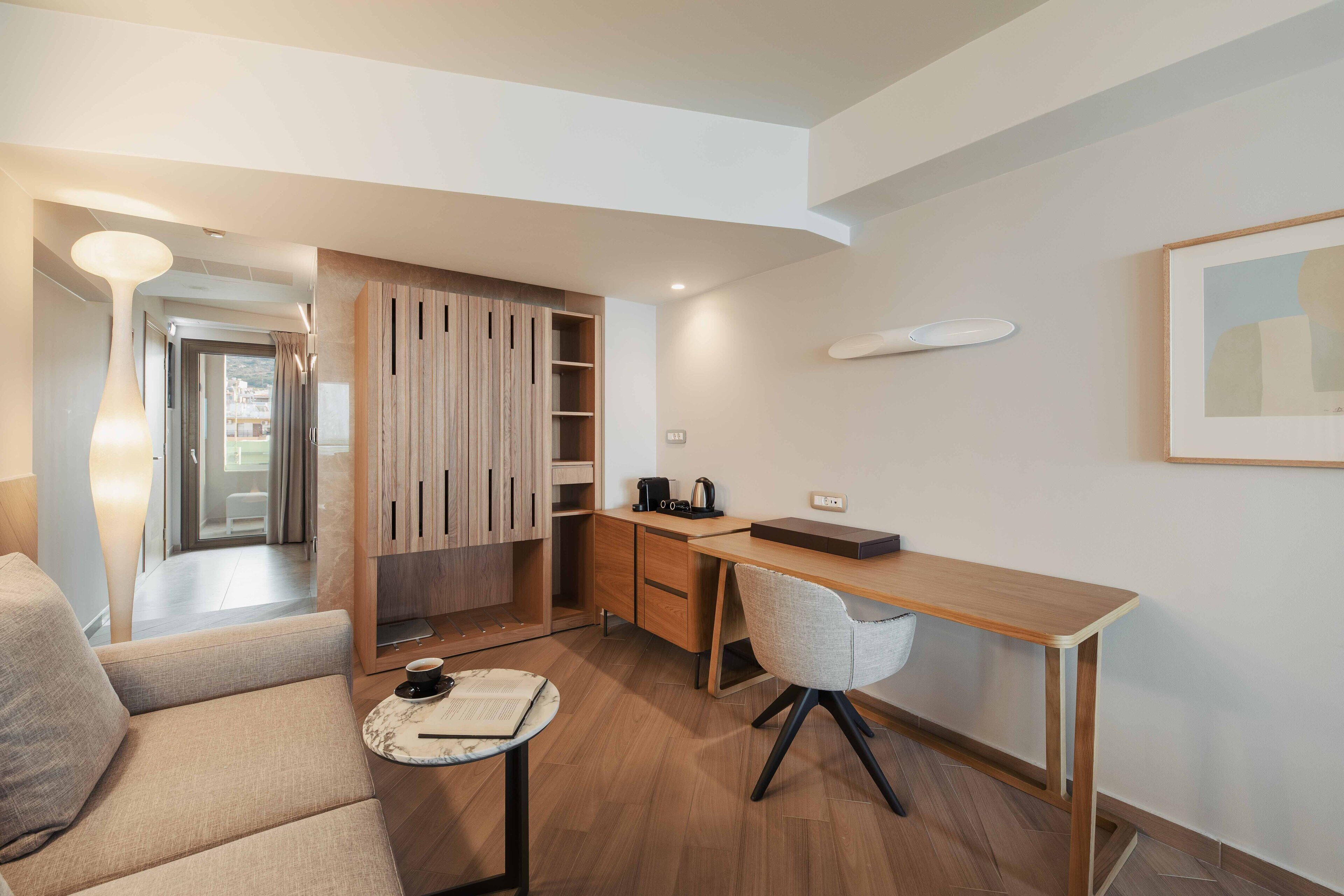 A modern hotel room with a wooden desk, a comfortable sofa, and a small round table with a book and a cup.