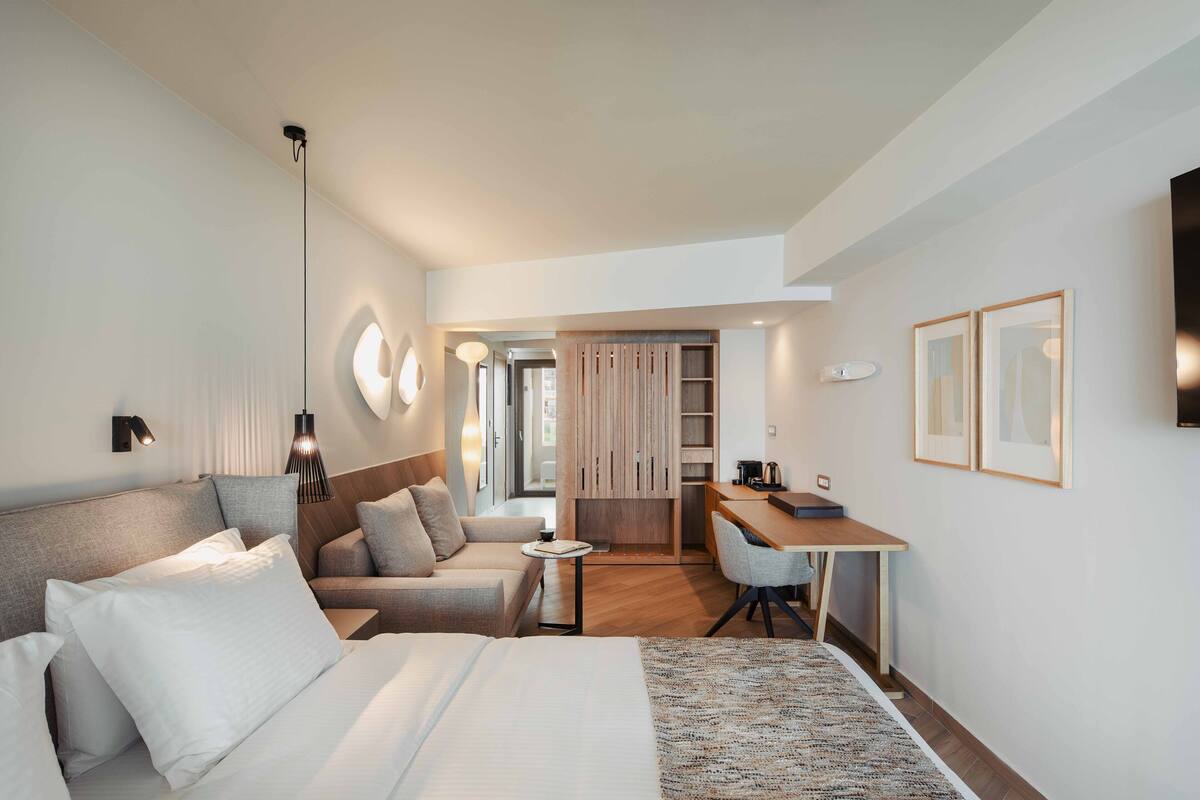 A modern hotel room with a large bed, a sofa, a desk, and a kitchenette.