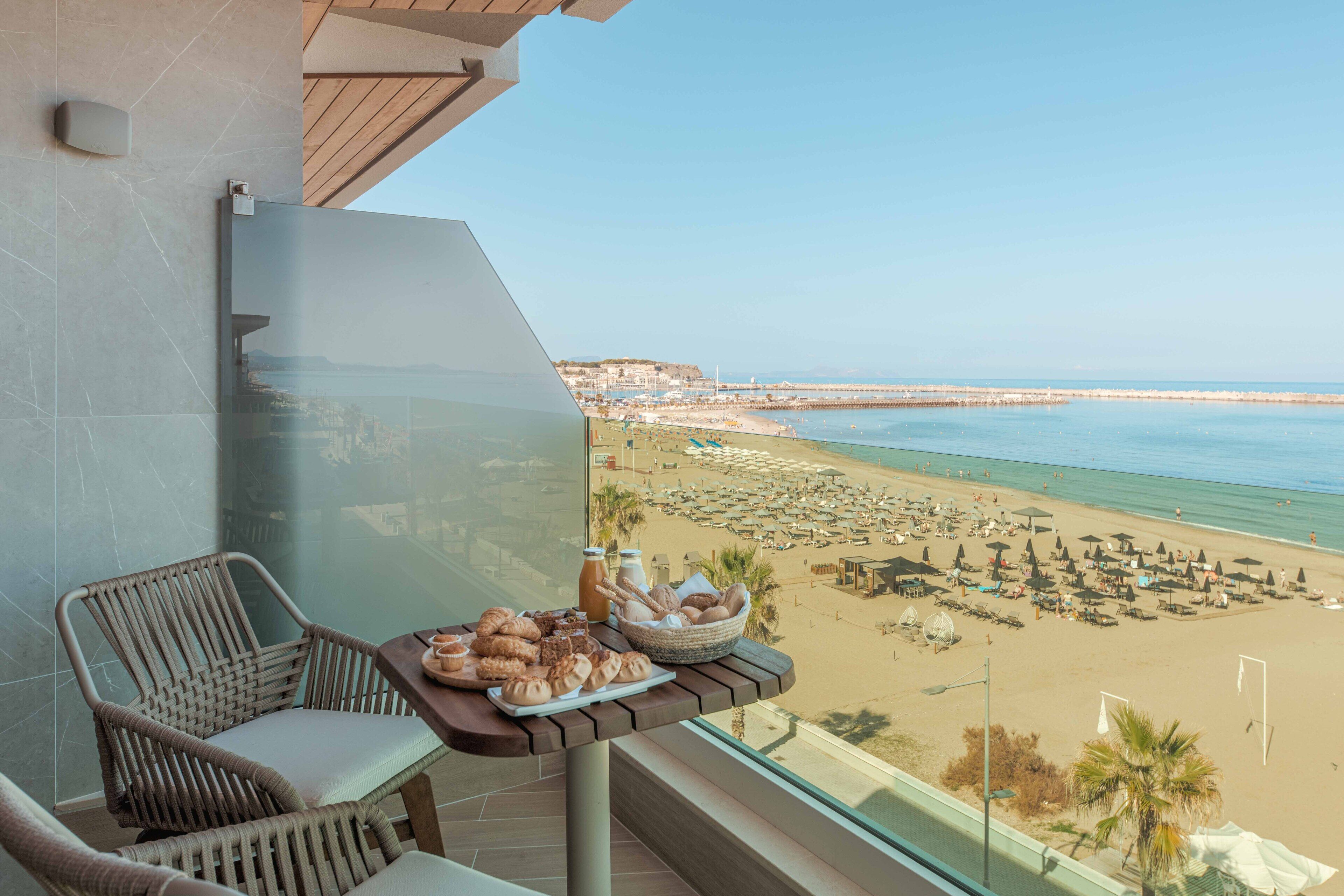 A balcony with wicker chairs, a table with pastries and drinks, and a view of a beach with umbrellas and people.