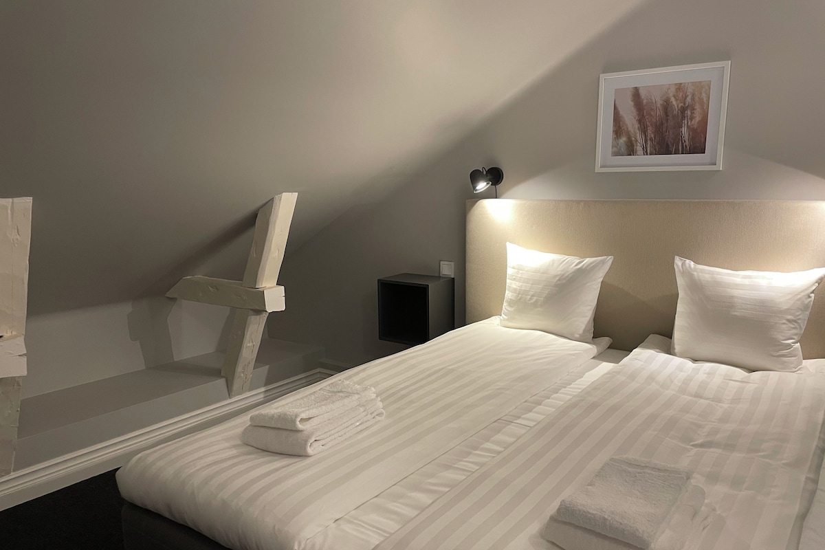 A neatly made bed with white linens and two pillows, a bedside table with a lamp, and a framed picture on the wall.