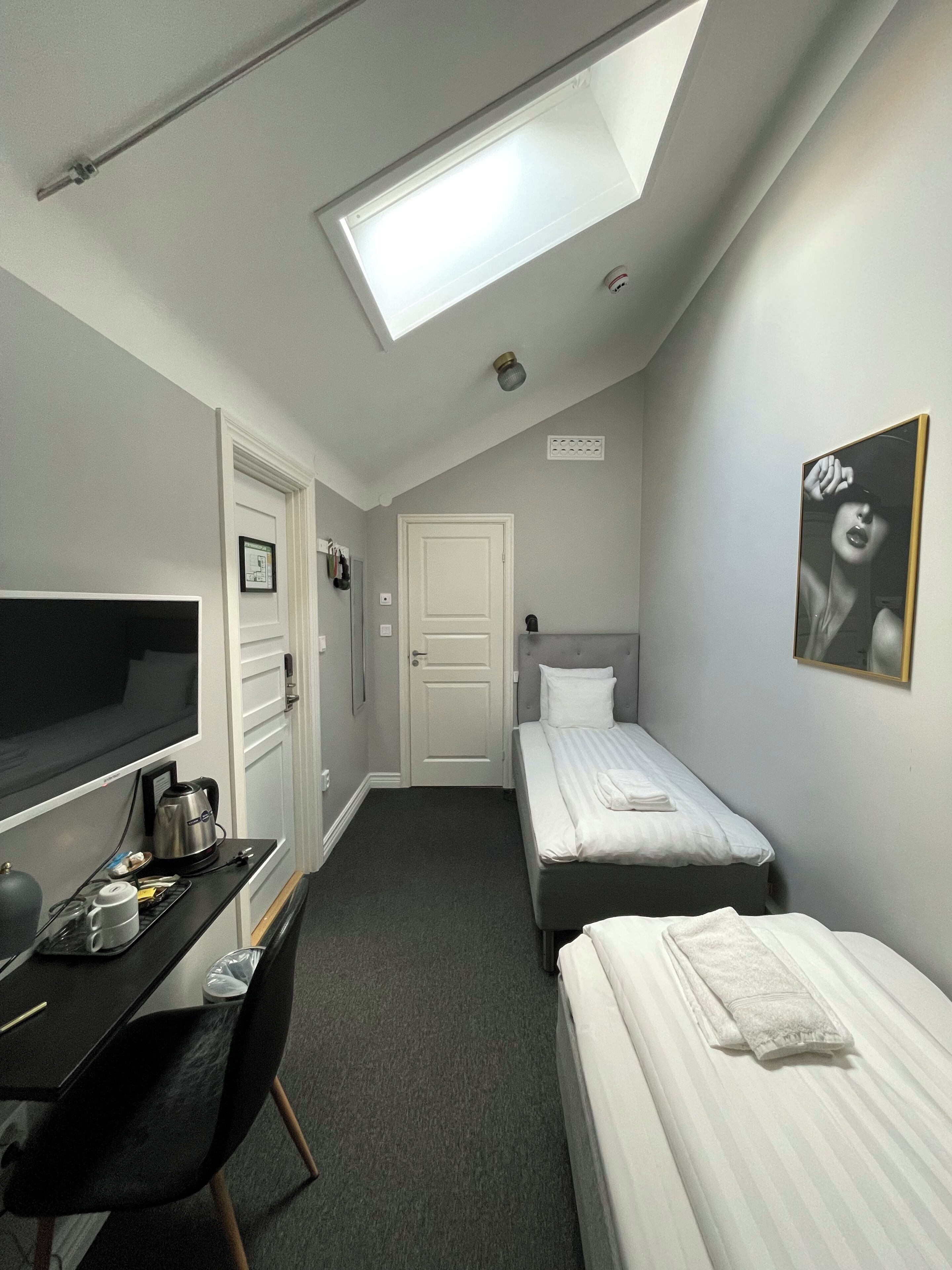 A compact hotel room with a skylight, a desk with a kettle, a TV, two beds, and a chair.