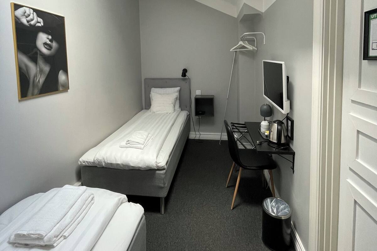 Habitación de hotel compacta con una cama individual, un escritorio con computadora, un bote de basura pequeño y una ventana que proporciona luz natural.