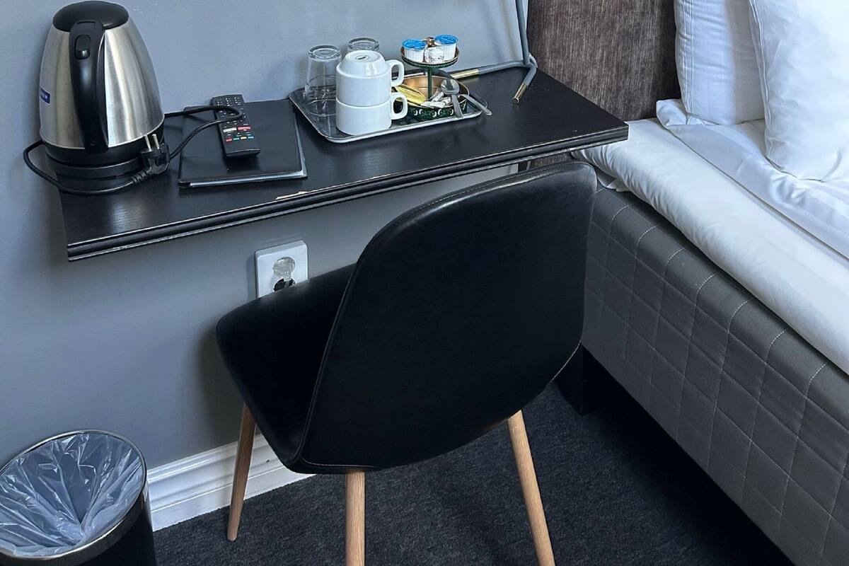 Habitación de hotel pequeña con cama, escritorio con hervidor, silla, lámpara y un cesto de basura.