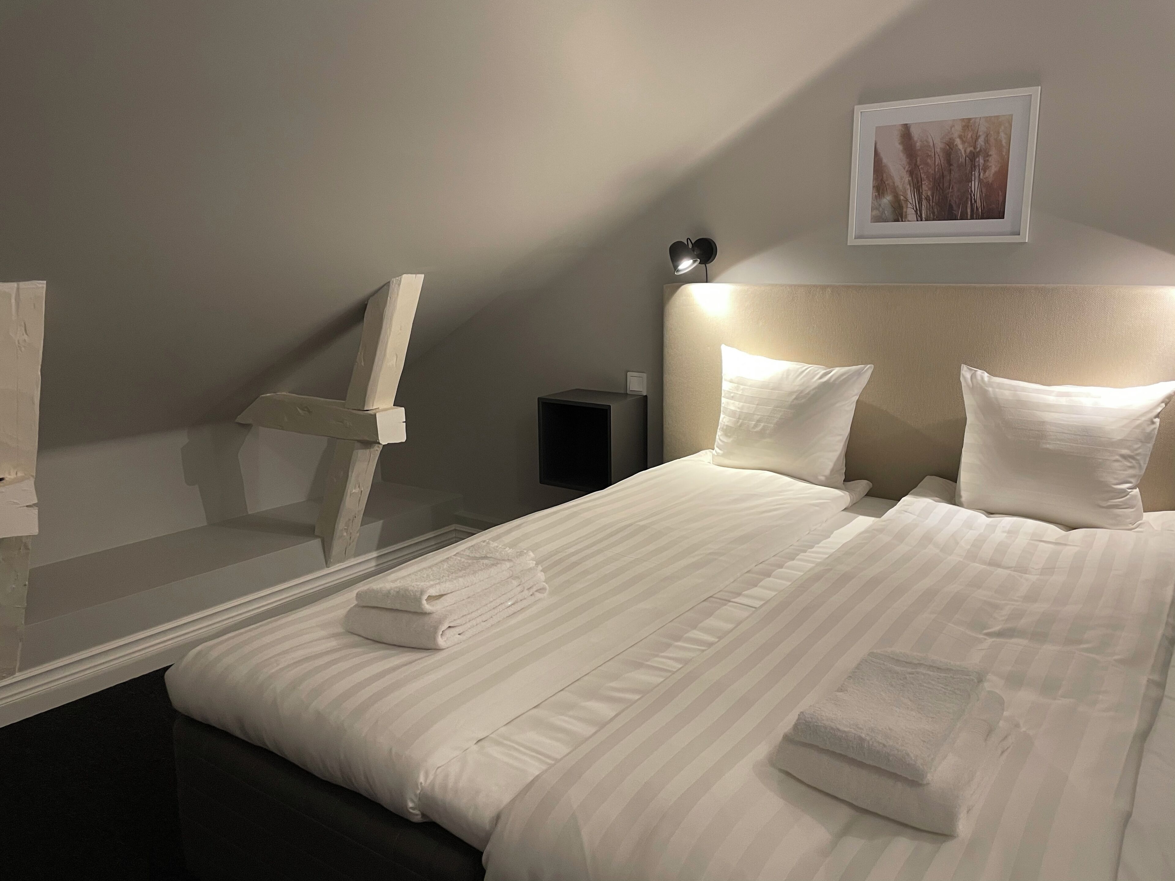 Cama bem arrumada com lençóis brancos, cabeceira com iluminação integrada, quadro na parede e mesa de cabeceira pequena com abajur.