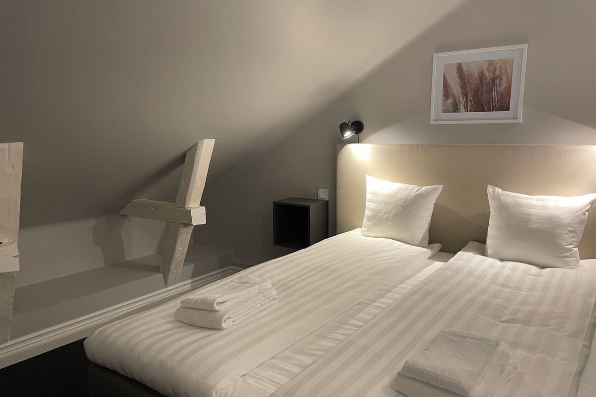 Una cama bien hecha con sábanas y almohadas blancas, una lámpara de noche y un cuadro en la pared.
