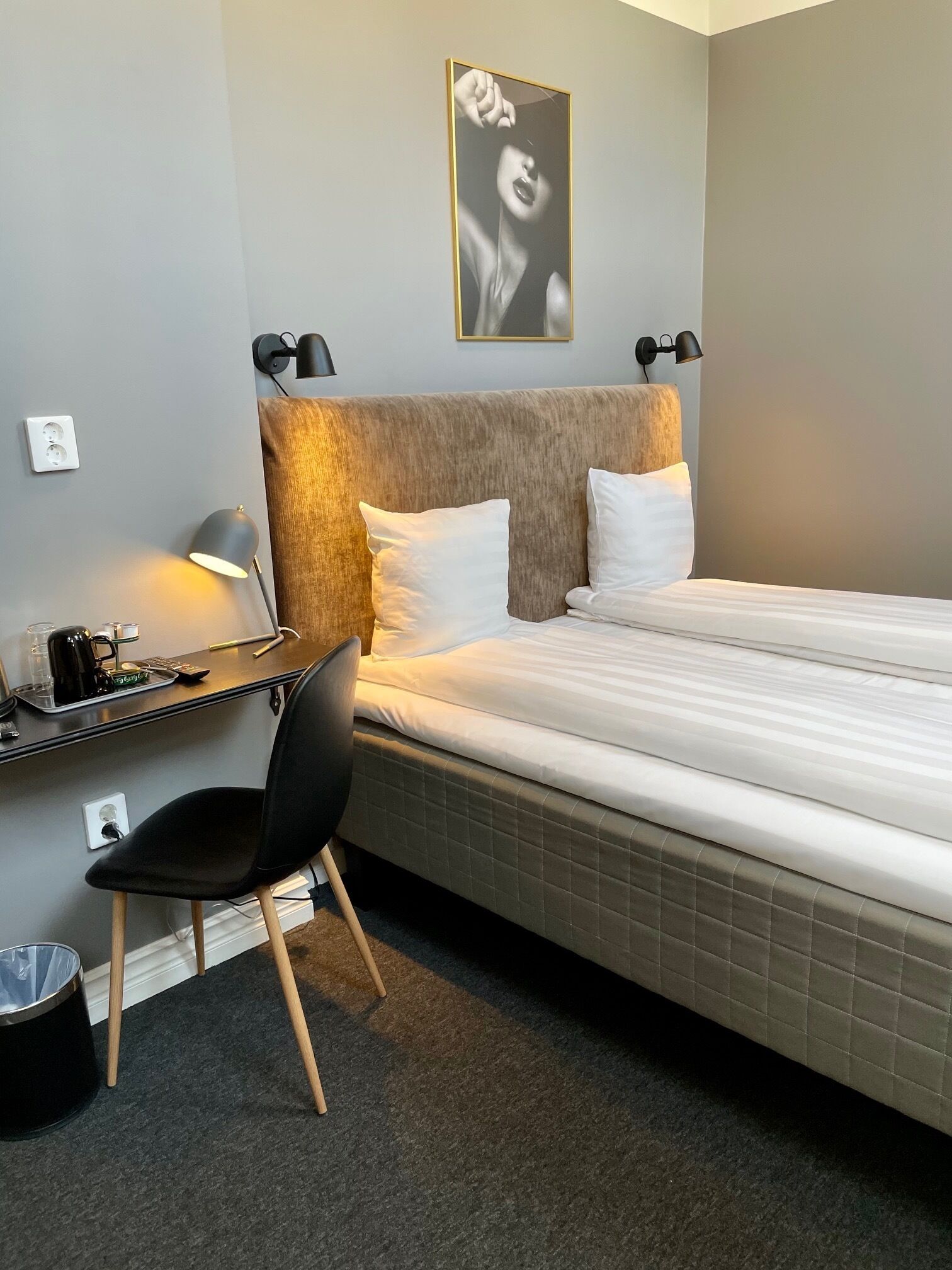 Quarto de hotel bem arrumado, com cama, escrivaninha, cadeira e luminária fixada na parede.
