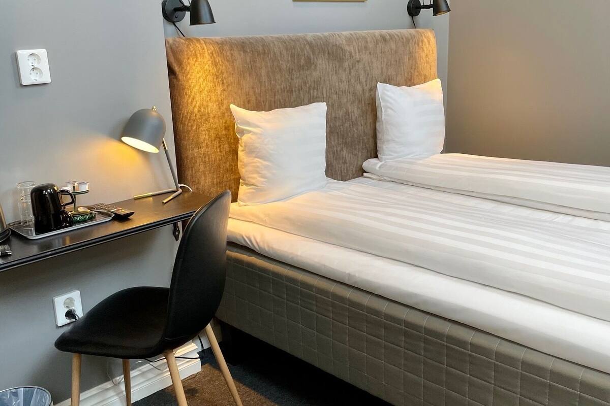 Habitación de hotel ordenada con una cama, un escritorio, una silla y una lámpara de pared.