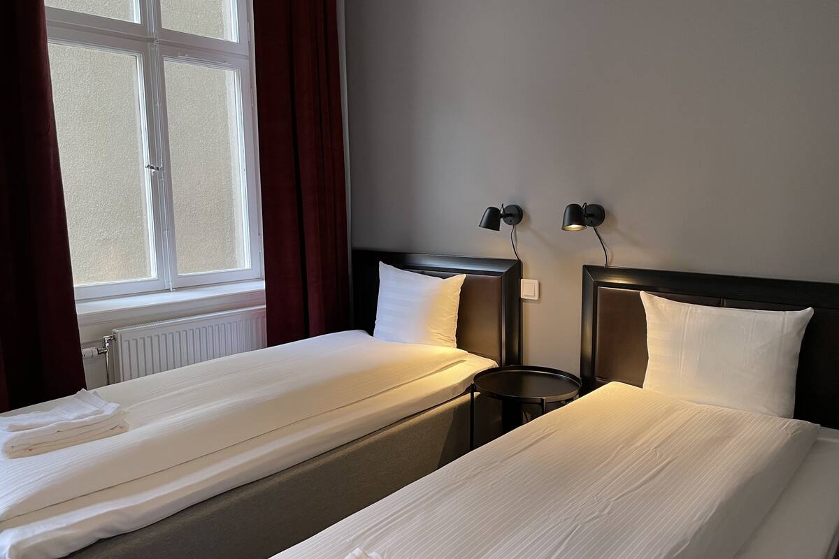 Habitación de hotel con dos camas individuales, una ventana con cortinas, una mesa redonda y dos lámparas fijadas a la pared.