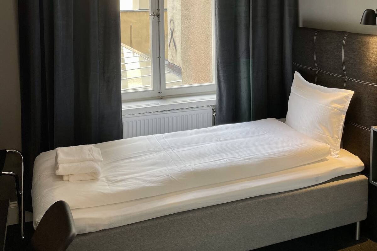Una habitación de hotel con cama, ventana con cortinas, radiador y cabecero.