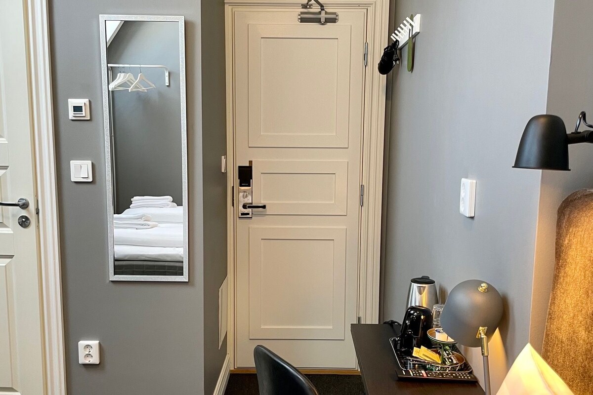 A hotel room with a bed, a desk with a lamp, a chair, a wardrobe, and a mirror.